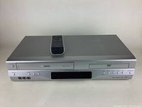 Front view of Toshiba DVD/VHS combo unit with remote on top, showing DVD tray and front panel controls and inputs.