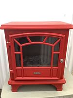 Front facing photo of red electric heater resembling a small stove with a segmented glass door and metal handle, placed on a table.