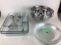 Photo showing nested Cuisinart stainless steel mixing bowls stacked on the right and several glass baking dishes stacked on the left including a pie plate with a green sticker on top, and three stainless steel utensils placed inside the smallest glass dish.