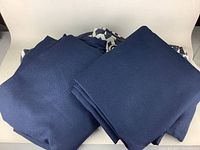 Two sets of folded drapes. One dark blue solid blackout curtains, the other partially visible darker patterned set.