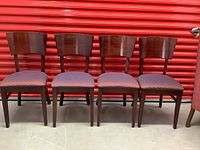 Front view of four wooden dining chairs with dark wood and upholstered seats, arranged side by side against a reddish backdrop.