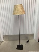 Floor lamp with woven natural shade, black slender metal stand, and square base, photographed against a corrugated wall and concrete floor.