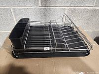 Clear view of the stainless steel dish rack showing the wire structure for drying dishes and the black plastic utensil holder on the left side. Base tray is black plastic and visible on all sides.