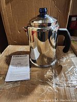 Faberware stainless steel 12 cup coffee percolator with black handle and blue knob on lid, alongside an instruction manual on cardboard background.