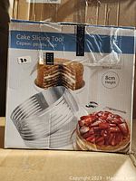Closed cardboard box with image and text describing a stainless steel cake slicing tool 8cm height, including visual of a sliced layered cake and a strawberry cake.