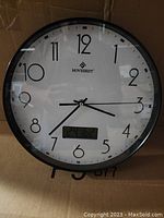 Front view of the Movebest analogue wall clock showing large black numbers, black hands, and a digital display near the bottom.