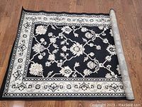Photo shows the folded rug partially unrolled on a wooden floor, displaying its black background with white floral design and bordered edges.