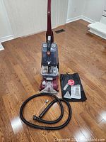 Full view of the Hoover Power Scrub carpet cleaner standing on wood floor with coiled black hose in front, small cleaning accessories, and manual alongside.