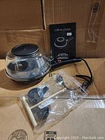 Curtis Stone electric skillet with glass lid, power cord, instruction manual, and wooden utensils displayed together on cardboard box.