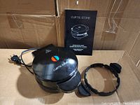 Photo of black Curtis Stone stuffed waffle maker, recipe book, and two detachable rings in cardboard box.
