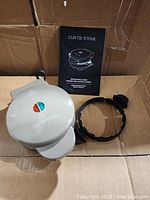Grey Curtis Stone waffle maker closed with detachable rings and instruction manual arranged beside it