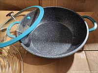 Curtis Stone 4.5QT cast aluminum pan with black speckled non-stick interior, teal exterior, glass lid, and metal rack inside cardboard box.