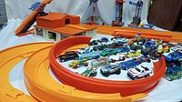 Full view of 1968 Mattel Hot Wheels Super Charger race track with orange track loops and ramps, base station, and 50 assorted Hot Wheels cars arranged inside loops and beside track sections.