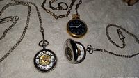 Three vintage pocket watches with attached chains laid on fabric. One shows open mechanical parts. One has black dial with gold-tone case and cracked glass. One hunter-case watch partially open showing interior workings.