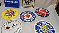 Overview showing 7 vintage metal garage signs including Chevrolet, Pontiac, Studebaker, Oldsmobile, Roaring Lion energy drink, and zombie parking
