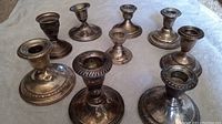 Nine assorted vintage sterling silver candlestick holders displayed on cloth surface. Each is unique in design, size, and style with visible tarnishing and some damage.