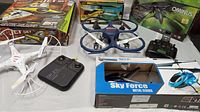 Photo showing collection of 5 remote-controlled flying vehicles including Syma drone, Air Hogs helicopter, boxed Sky Force helicopter, dark blue drone with guards, and Dromeda Ominus FPV drone in box with controller.