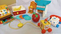 Photo shows a collection of vintage Fisher-Price toys including a yellow and orange cash register, music box record player with records, lift and load depot, wobble apple, telephone pull toy, and squeeze pull pop-up toy.