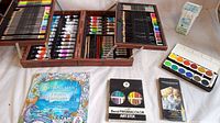 Wooden box open showing compartments filled with colored pencils, paint markers, art sticks, and a watercolor palette. Also shows coloring books and art tools placed near the box.
