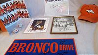 Wide shot showing autographed Chicago Bears photo, autographed vintage Broncos framed photo, orange signed Broncos cap, signed baseballs, and a signed blue 'BRONCO DRIVE' sign on table.