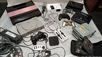 Wide view of all video game consoles and accessories arranged on a white surface, including Atari 5200, PlayStation, Sega Genesis, Xbox, Nintendo Wii, various controllers, and cables.