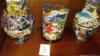 Three glass jars filled with assorted vintage costume jewelry items such as necklaces, bracelets, rings, earrings, and beads shown from the front on a wooden surface.