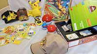 Wide view of Pokemon collectibles including hats, plush, handheld game, cards laid out, game board box, Pokeball containers and walkie-talkie half.