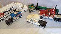 Wide photo showing all five semi trucks arranged in a row on a gray surface. From left to right: blue semi truck, white Target semi truck with trailer, small green Timberland Express semi cab with trailer, red Tonka hook and ladder fire truck semi cab, white Tiara yachts semi cab.