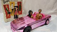 Photo showing metallic pink Mattel Corvette toy car from 1984 with two black dolls seated inside, male Ken doll 1968 and black Barbie 1966. Barbie doll case visible in background.