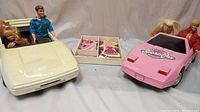Front view of two sports car toys, white and pink, with Barbie and Ken dolls seated. Magnetic dress-up set placed between the two cars.