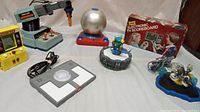Overview photo showing all items: Radio Shack Armitron drill toy, Tyco Doodle Dome, Radio Shack Electronic TV Scoreboard game in box, Pac-Man handheld game, Activision and Lego Portal of Power bases, two Skylanders figures.