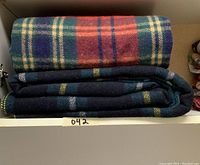 Two folded wool blankets stacked in a shelf. Top has red, green, blue, and beige tartan plaid. Bottom blanket is dark green with blue and yellow plaid lines.