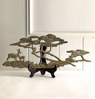 Brass wall hanging displayed on a black wooden stand showing deer, grass, and stylized tree branches.