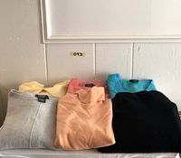 Six sweaters including pastel yellow cardigan, peach turtleneck, coral crew neck, blue-green V-neck, light gray and black pullovers, laid out folded against white wood paneling.