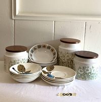 Three white Hornsea ceramic canisters with wood lids and green floral pattern, front view with three stacks of dishes: six shallow bowls with fruit motif and six soup bowls with geometric floral border.