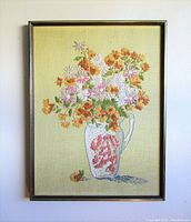 Full front view of framed embroidery showing colorful floral bouquet in a pitcher on yellowish fabric background.