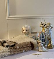 Photo shows a group of ceramic and decorative figurines including a poodle planter, ceramic lady with parasol, paper mache cat, Dutch house ornament and brass bell, placed on a white surface.