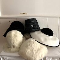 Full front view of five hats showcasing variety of materials: plush white fur, black velvet and straw. Hats arranged on white surface against white paneled wall.