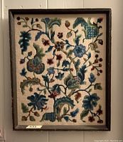 Front view of framed needlepoint artwork showing Jacobean floral pattern with blue and green threads in a wooden frame.