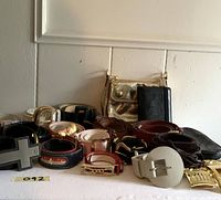View of multiple belts rolled or folded in various colors and designs, belt buckles and a leather gold poppy bag placed on a flat surface.