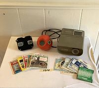Front-facing view of the lot showing the black View-Master viewer, orange spherical finder, gray GAF projector, and gray Panasonic radio on a white surface with various stereo reels scattered around.