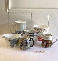 Group shot of 6 vintage ceramic pieces including jugs, creamers, and bowls with floral and patterned designs on a white surface against a white paneled wall.