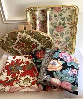 Three rectangular metal serving trays with cream backgrounds and vintage floral patterns, plus one oval serving tray with a tapestry floral pattern. Multiple pieces of vintage floral drapery fabric with different colors and patterns are arranged in front.
