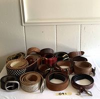 Photo showing multiple rolled vintage leather belts placed horizontally on a white surface against a white wall, featuring plain and tooled designs with different buckles.
