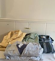 Four men's pajama sets laid out on a surface against a wall, showing various colors and patterns including beige, green geometric, dark blue and gray stripes, and white with blue stripes