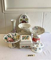 Front view of nine vintage pottery pieces including hand painted porcelain vase, pedestal dishes, ceramic powder box, candlesticks, and wall plaque. Items arranged in front of a white wall and shelf.