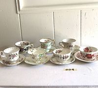 Seven vintage tea cups and saucers displayed side by side showing floral patterns and gold rims, arranged in two rows on white surface.