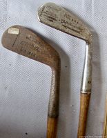 Photo showing close-up of two antique golf clubs, one labeled Champion A. McArthur Mid Iron and the other Spalding Elite, both with aged metal club heads and hickory shafts.