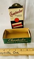 Front view of vintage Chantecler cigarette papers dispenser with lithographed design and MaxSold tag for lot 30.