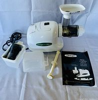 White Omega juicer machine with its components including a pulp collector, plunger, cleaning brush in holder, and instruction manual on a white background.